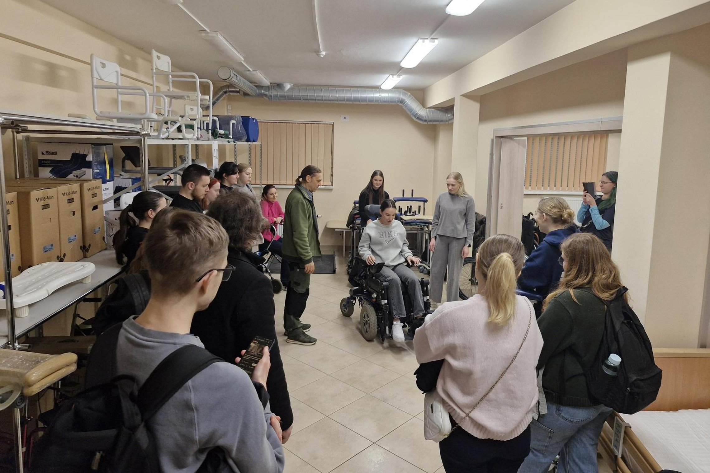 Vilniaus kolegijos studentai stebi, kaip jų kolegė valdo elektrinį vežimėlį valdymo pultelio pagalba.