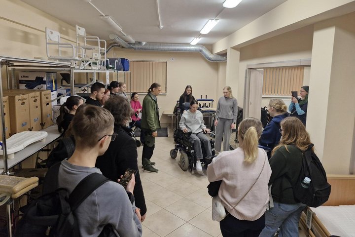 Vilniaus kolegijos studentai stebi, kaip jų kolegė valdo elektrinį vežimėlį valdymo pultelio pagalba.